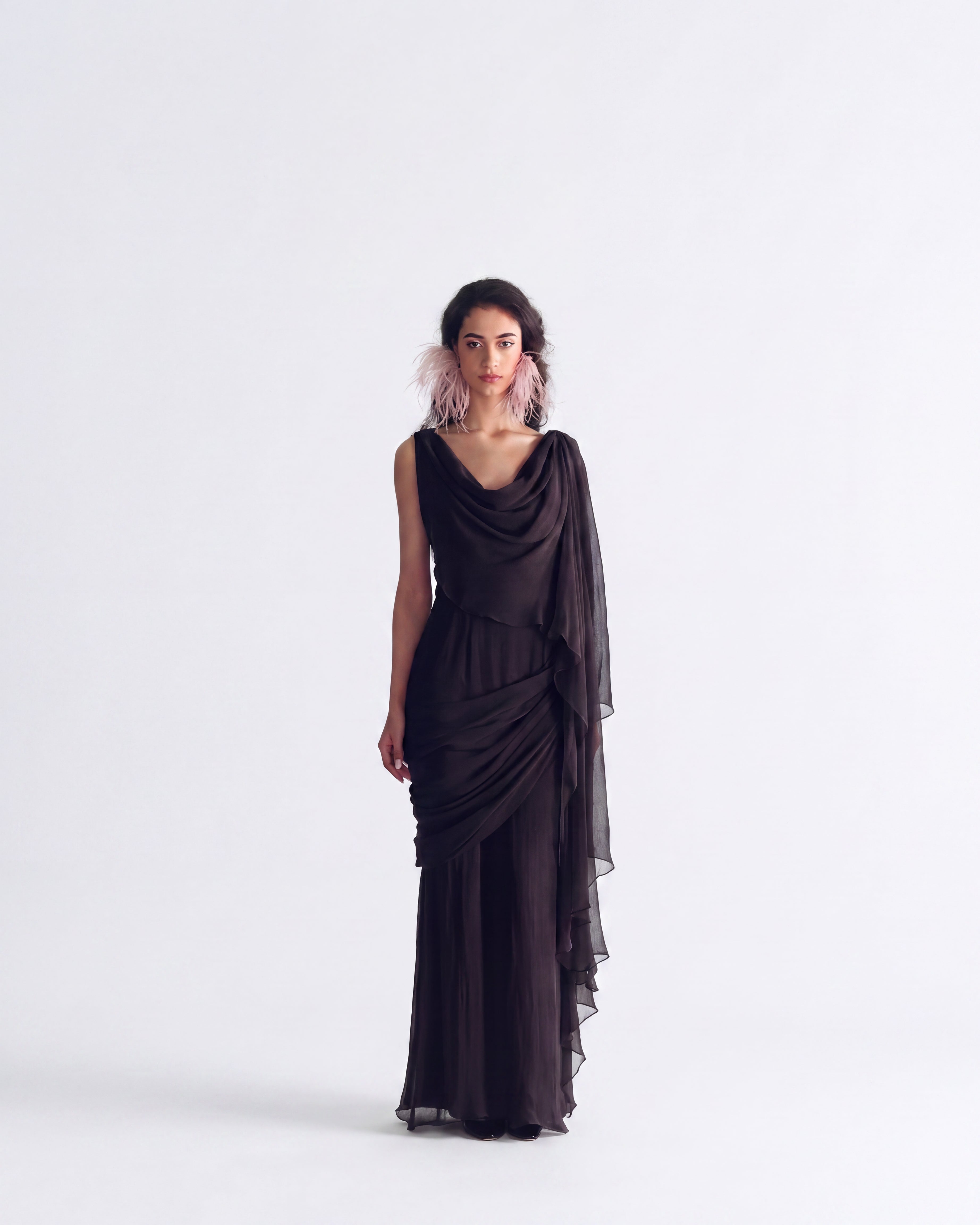 A model wearing a floor-length, draped black gown with a cowl neckline and a sheer cascading overlay, standing against a seamless light gray studio background.