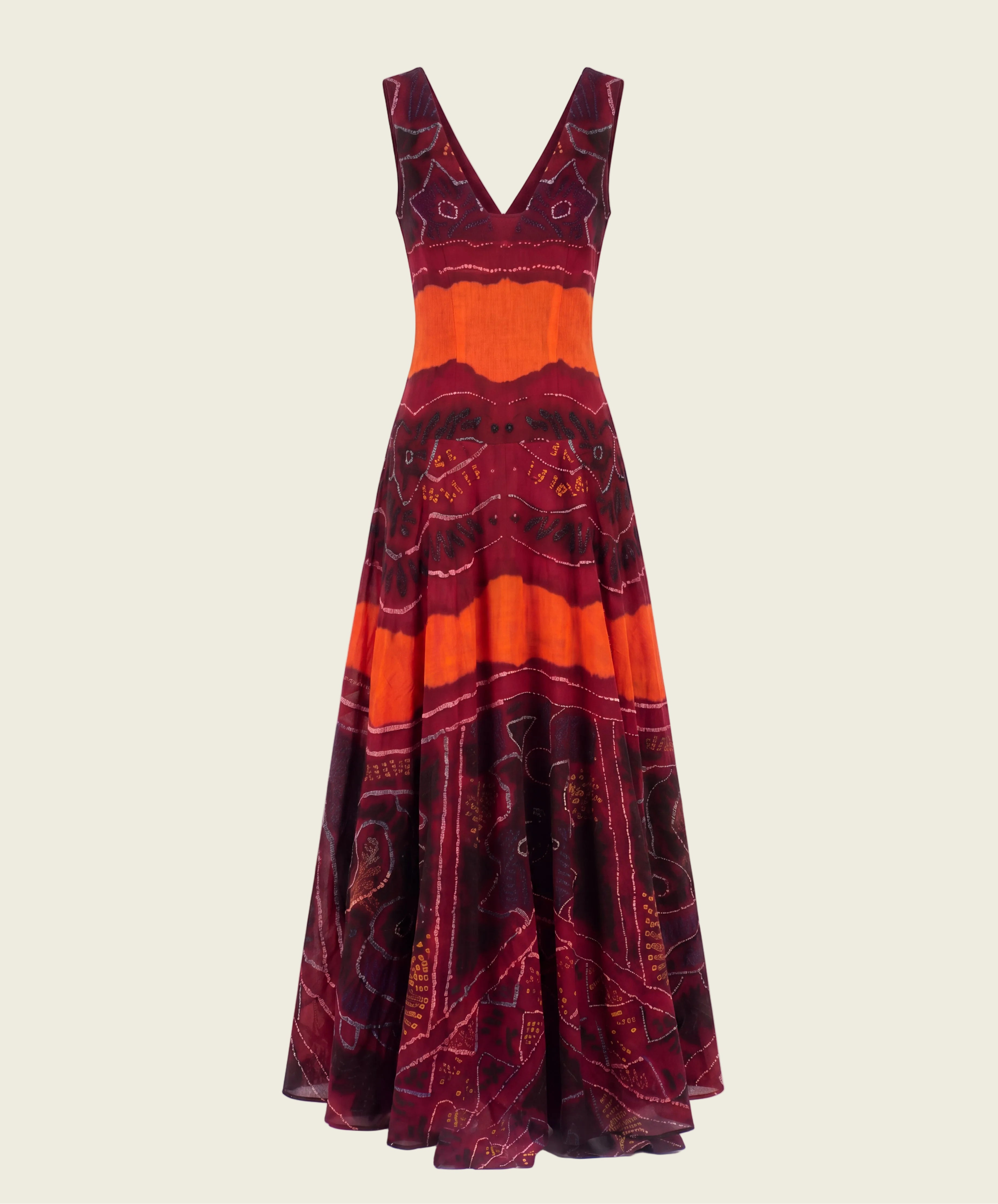 A sleeveless maxi dress with a deep V-neckline, featuring a bold pattern of orange, burgundy, and black with gold accents, displayed against a white background with a faux brick wall.