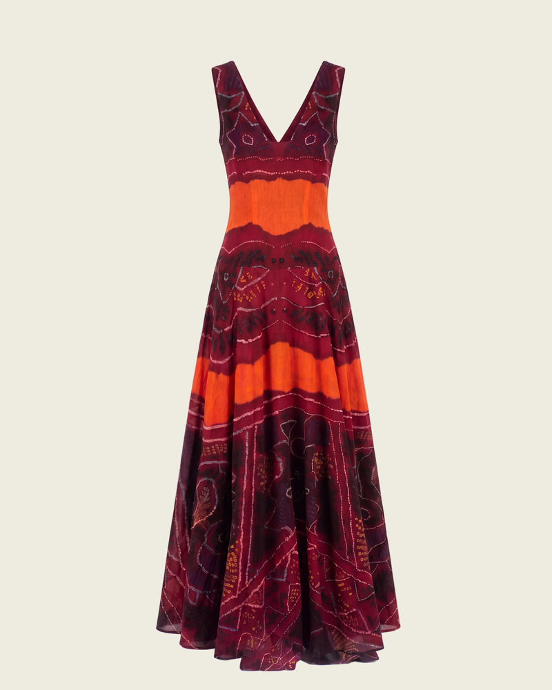 A sleeveless maxi dress with a deep V-neckline, featuring a bold pattern of orange, burgundy, and black with gold accents, displayed against a white background with a faux brick wall.